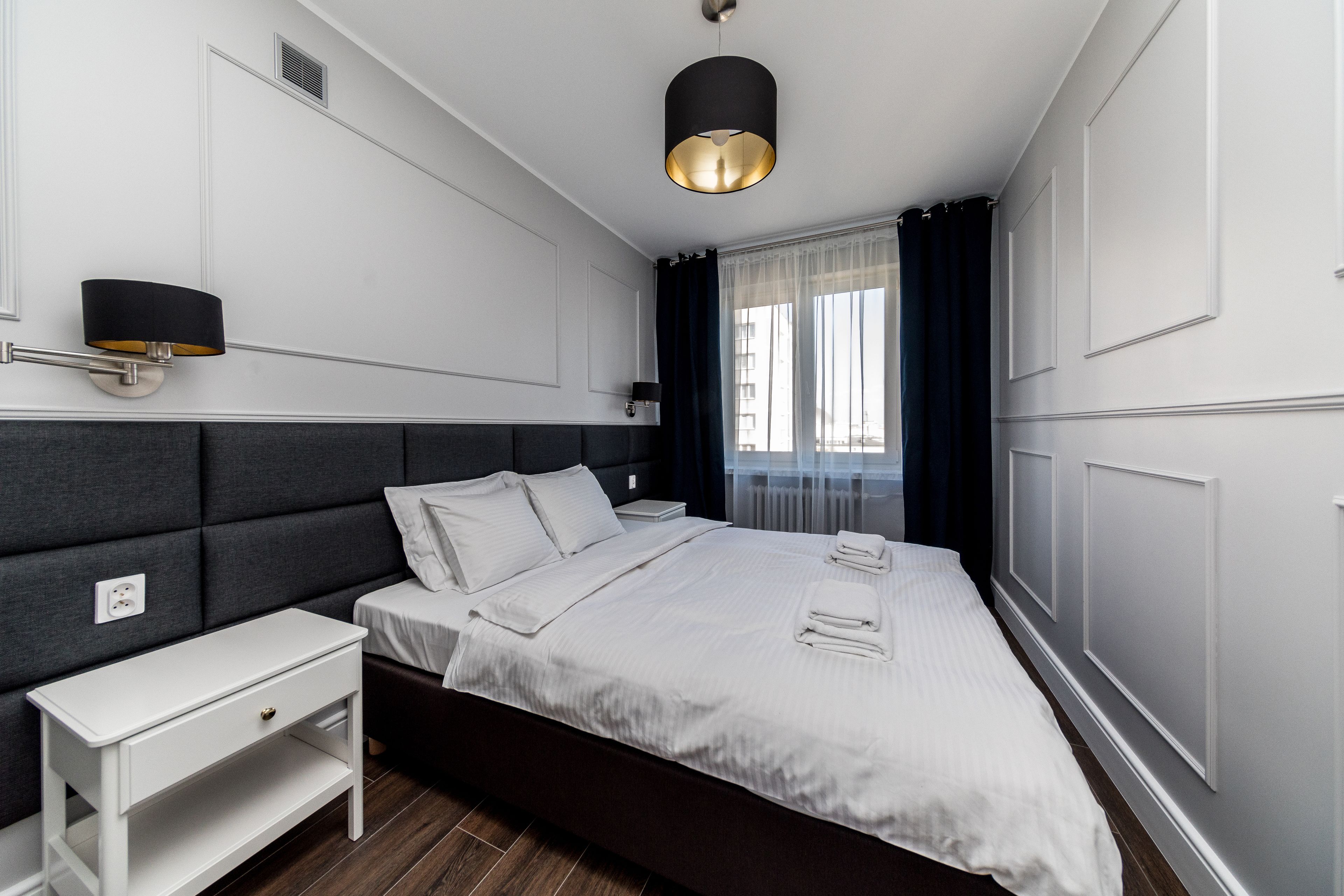 A modern hotel room with a large bed, a bedside table, a wall-mounted lamp, and a window with curtains.