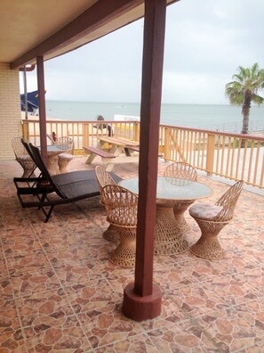 A covered outdoor seating area with wicker furniture and a stone-tiled floor, overlooking the beach and ocean. - Capri Beach Hotel (Corpus Christi)