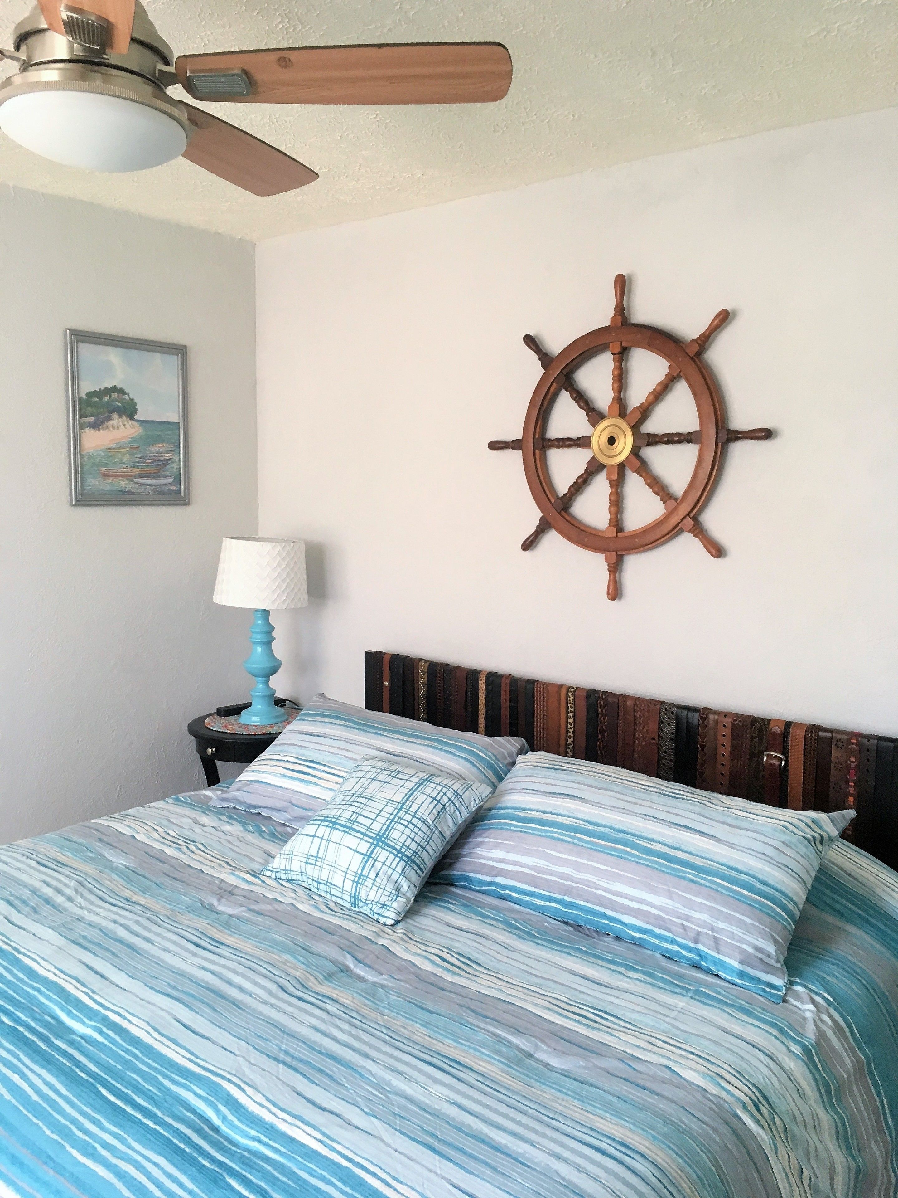 A bedroom with a bed, a wooden ship's wheel wall decoration, a small table with a lamp, and a framed picture on the wall.
