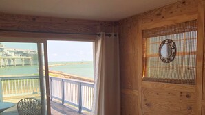 Una habitación con un gran ventanal corredizo que da a un balcón con vistas a una playa y un edificio. Hay una pared de madera con un reloj y un pequeño banco.