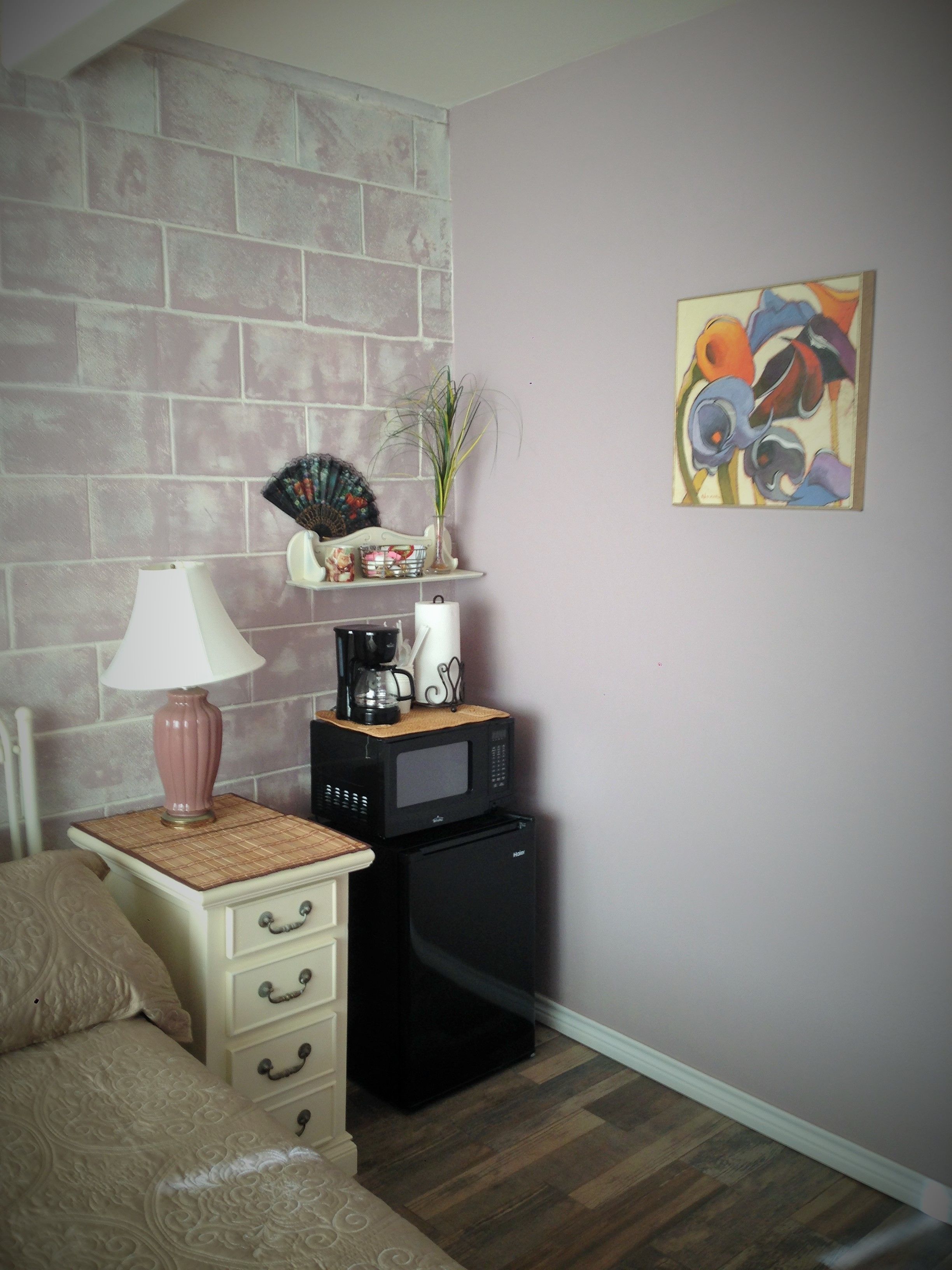 A bedroom with a bed, a bedside table with a lamp, a coffee maker, a microwave, and a refrigerator. There is a decorative fan and a painting on the wall.
