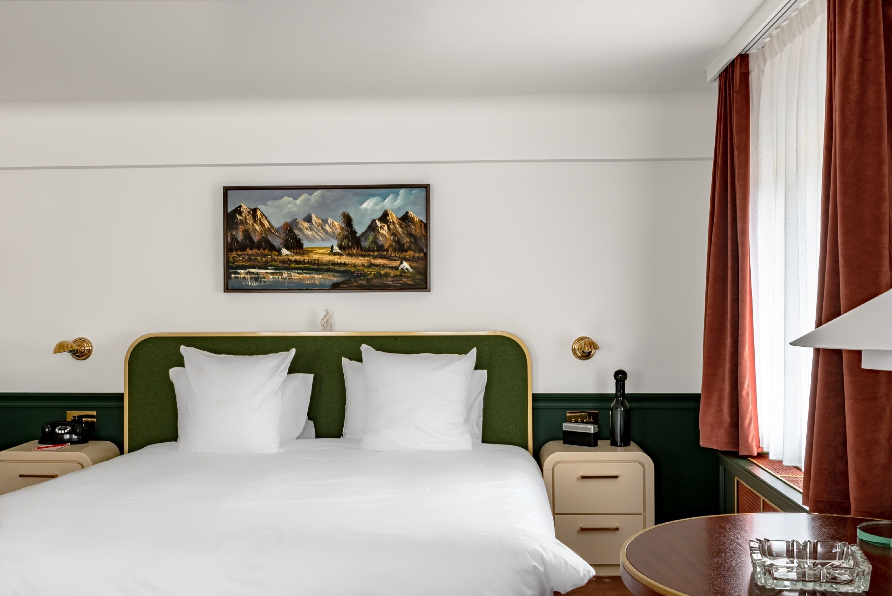A hotel room with a bed, a painting of mountains, a nightstand, a lamp, and a window with curtains.