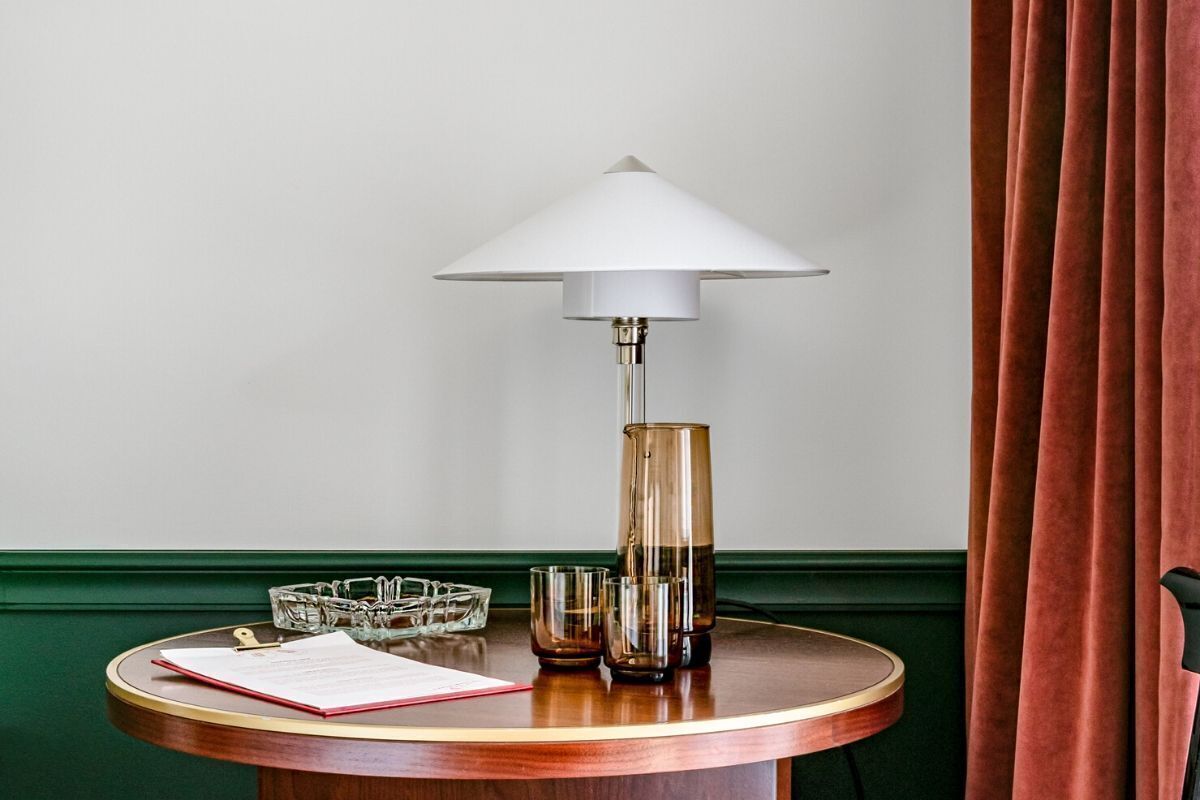 Een ronde houten tafel met een witte lampenkap, een glazen karaf en een set glazen.