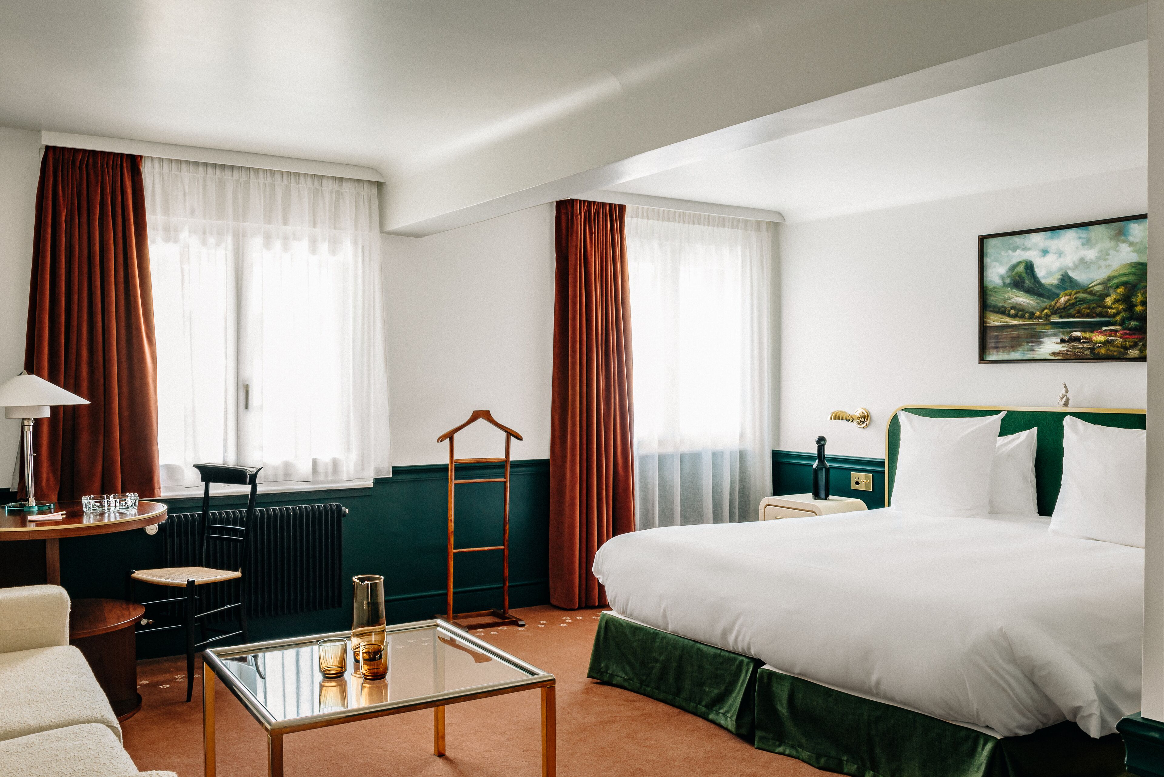 Habitación de hotel con una cama grande, un escritorio con lámpara, una silla, una mesa con vasos y un cuadro en la pared.