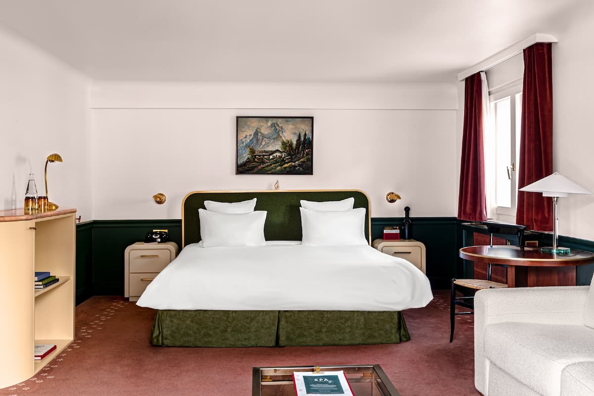 A hotel room with a large bed, a desk, a chair, a bookshelf, and a painting on the wall.