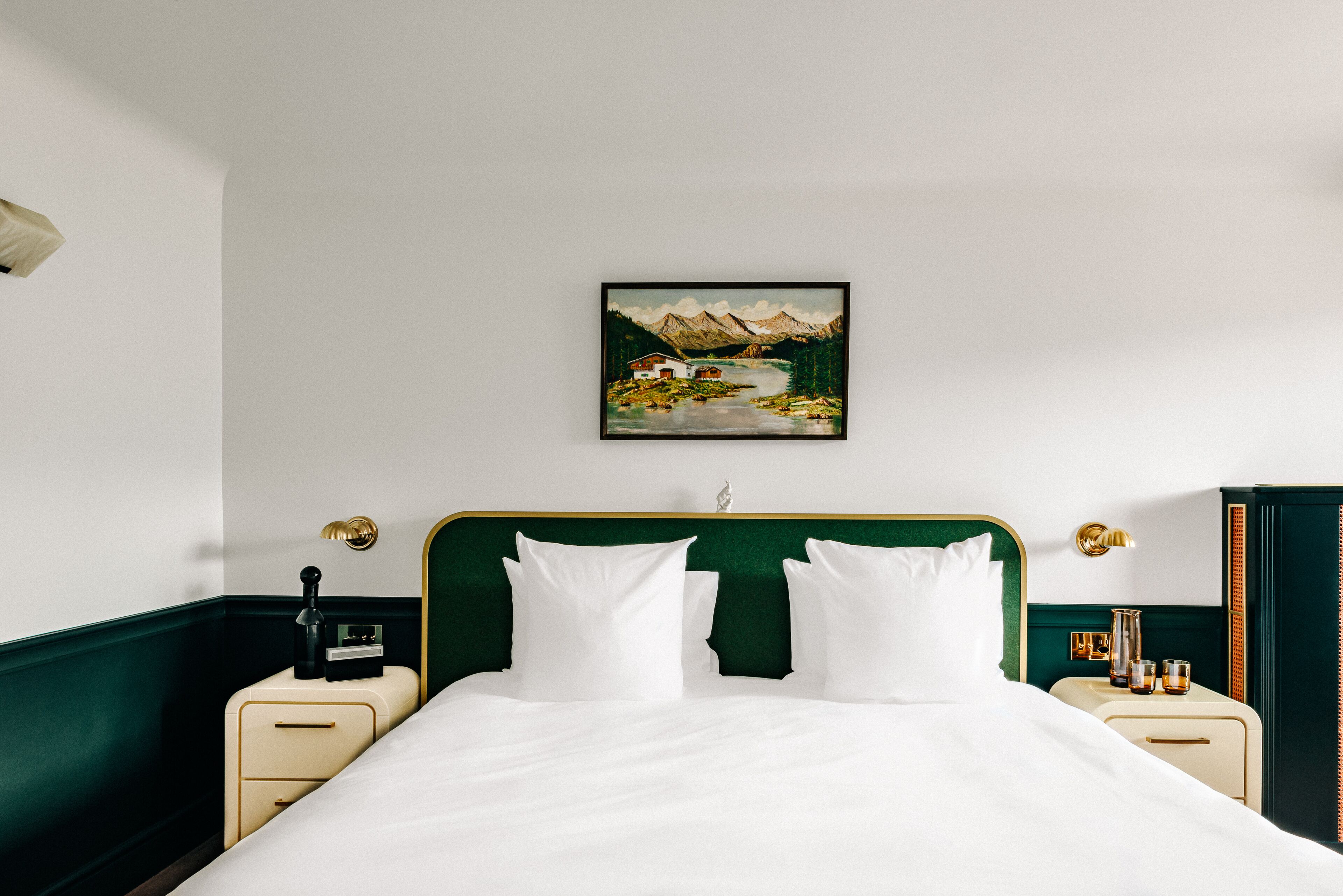 A neatly made bed with white linens, a green upholstered headboard, and a painting of a mountain landscape on the wall.