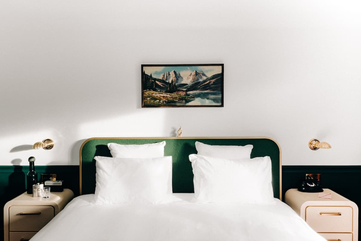 A neatly made bed with white linens, a green headboard, and a painting of mountains and a lake above it.