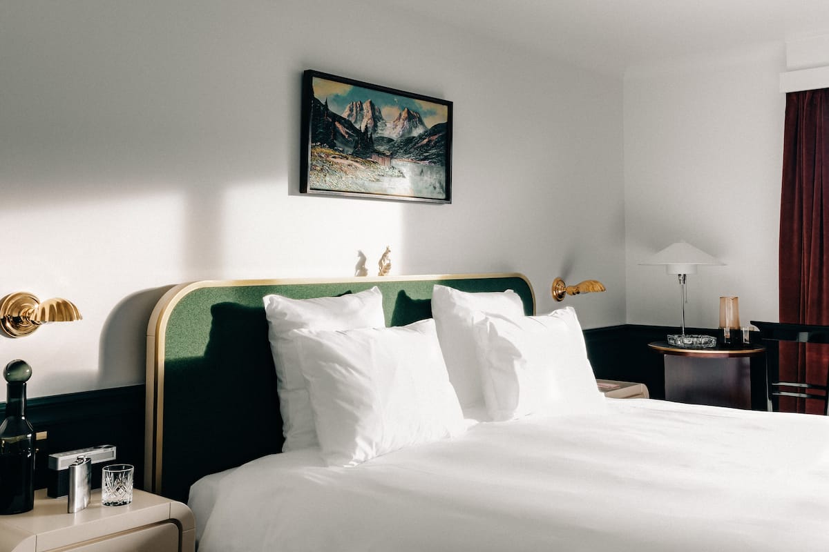 A neatly made bed with white linens, a framed painting of a mountain landscape on the wall, and a bedside table with a lamp and a small decorative item.