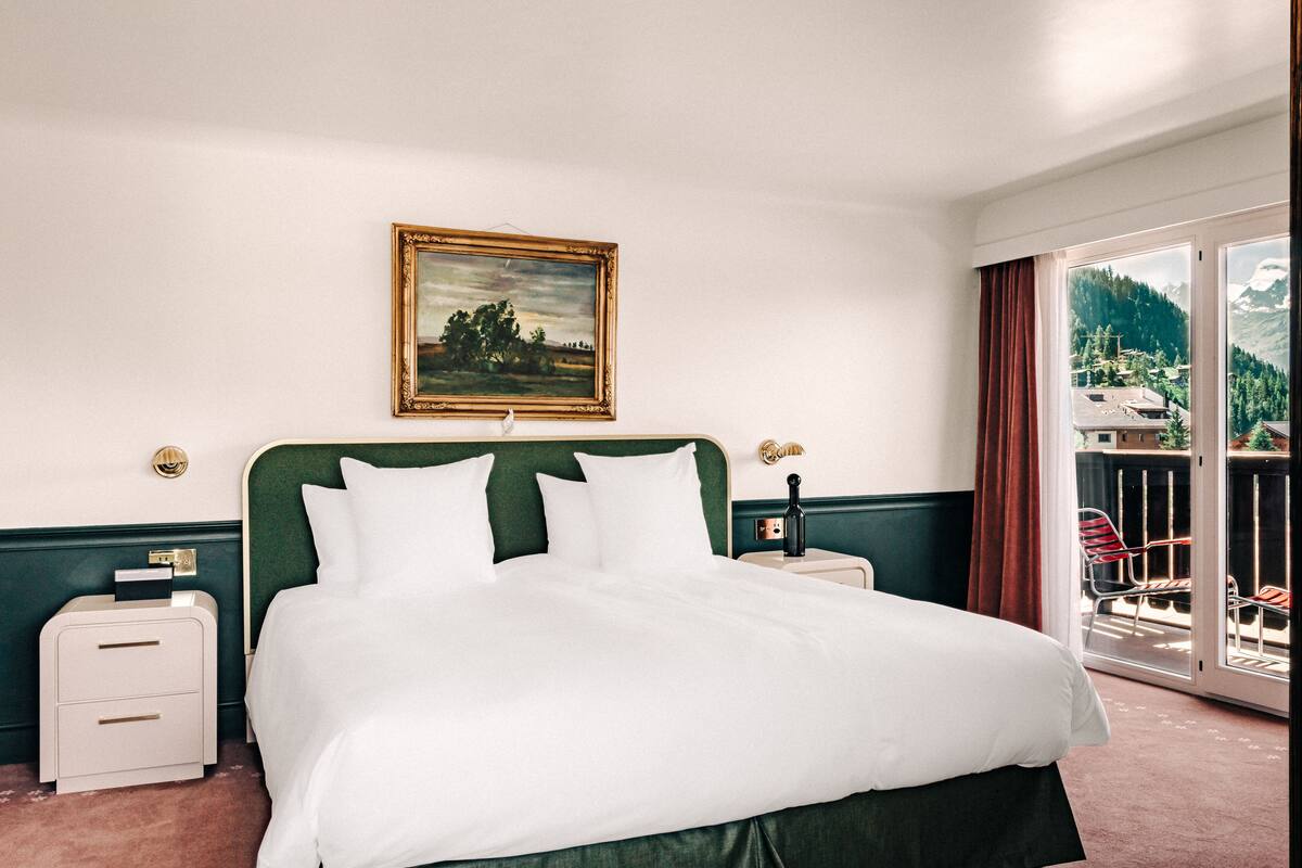 A hotel room with a large bed, two bedside tables, a painting on the wall, and a balcony with a view of the mountains.