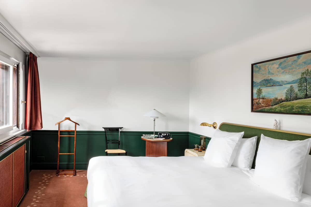 A hotel room with a bed, a desk with a lamp, a chair, a coat rack, and a painting on the wall.