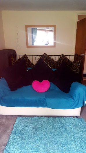Ein Bett mit blauer Bettdecke, schwarzen und gemusterten Kissen sowie einem pinkfarbenen herzförmigen Kissen und einem blauen Teppich.