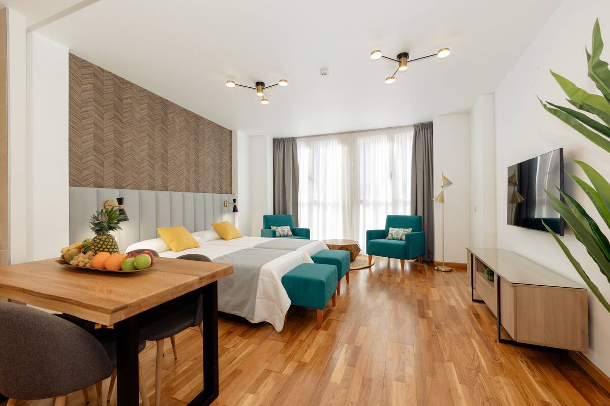 Een moderne hotelkamer met een houten vloer, een bed met grijs en wit dekbed, teal fauteuils, een houten nachtkastje met fruit, en een televisie aan de muur.