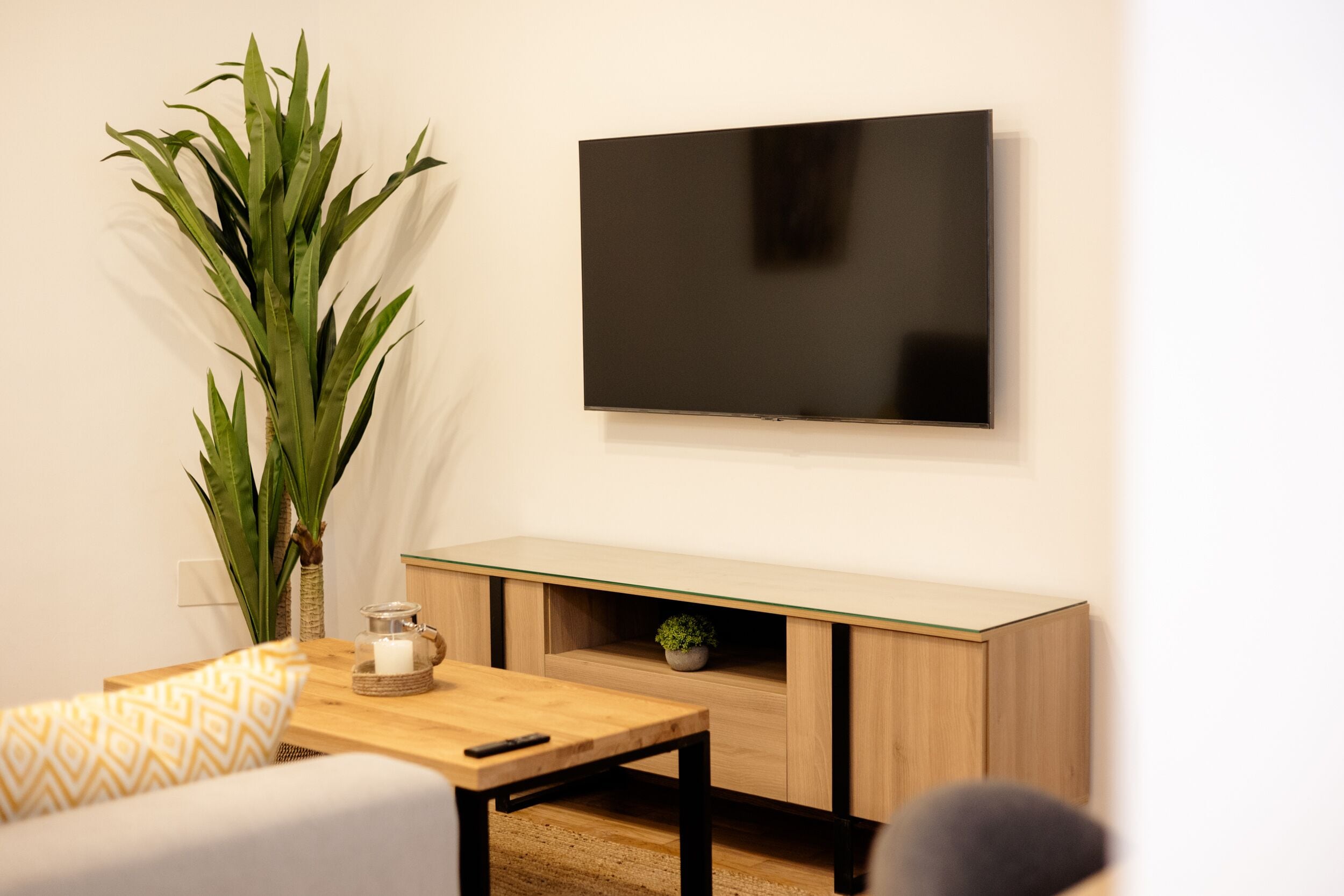 Una sala de estar moderna con un televisor de pantalla plana montado en la pared, una mesa de centro de madera, un sofá con cojines estampados y una planta verde grande.