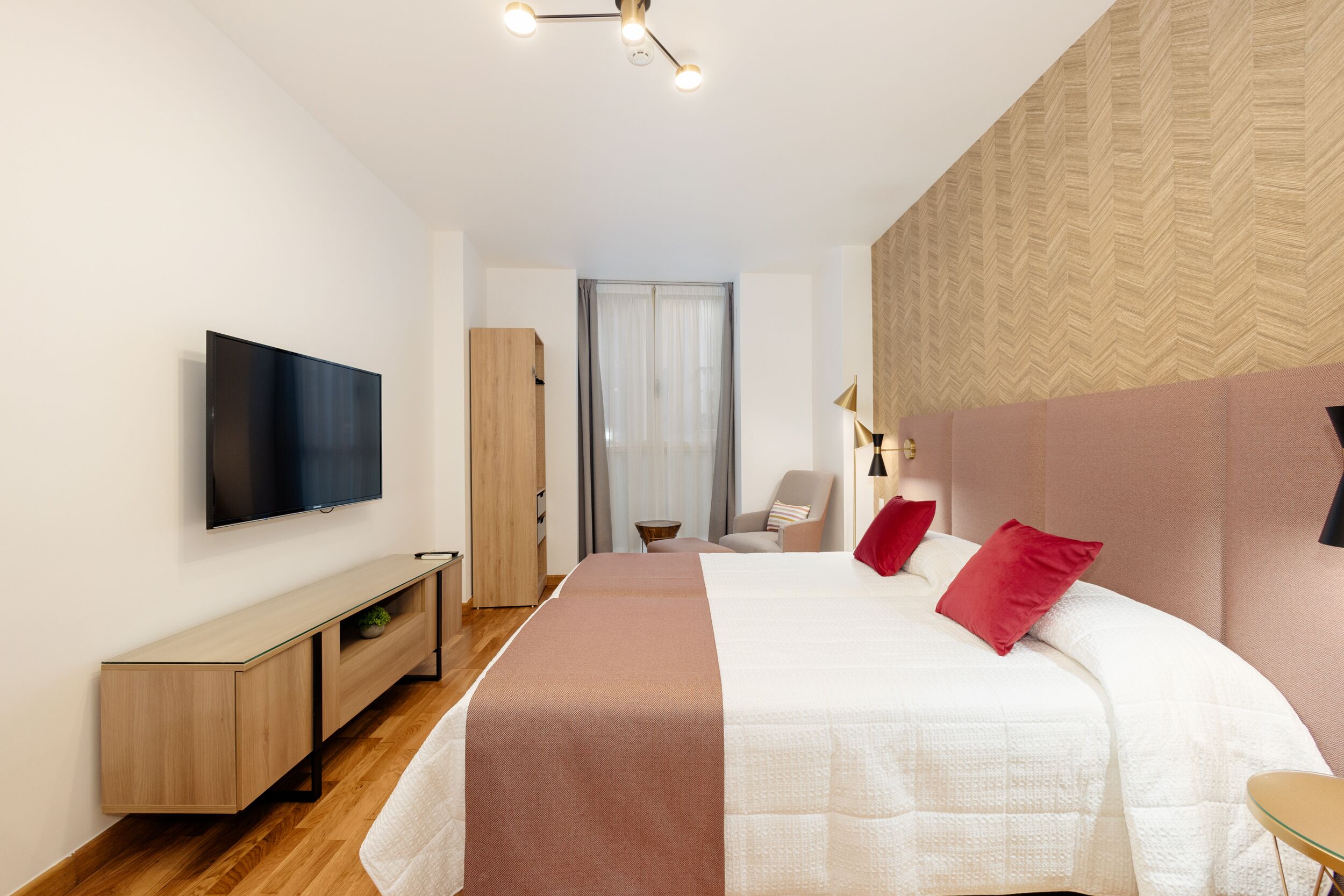 A modern hotel room with a large bed, a flat-screen TV, a wooden console table, and a view of the city through a window.