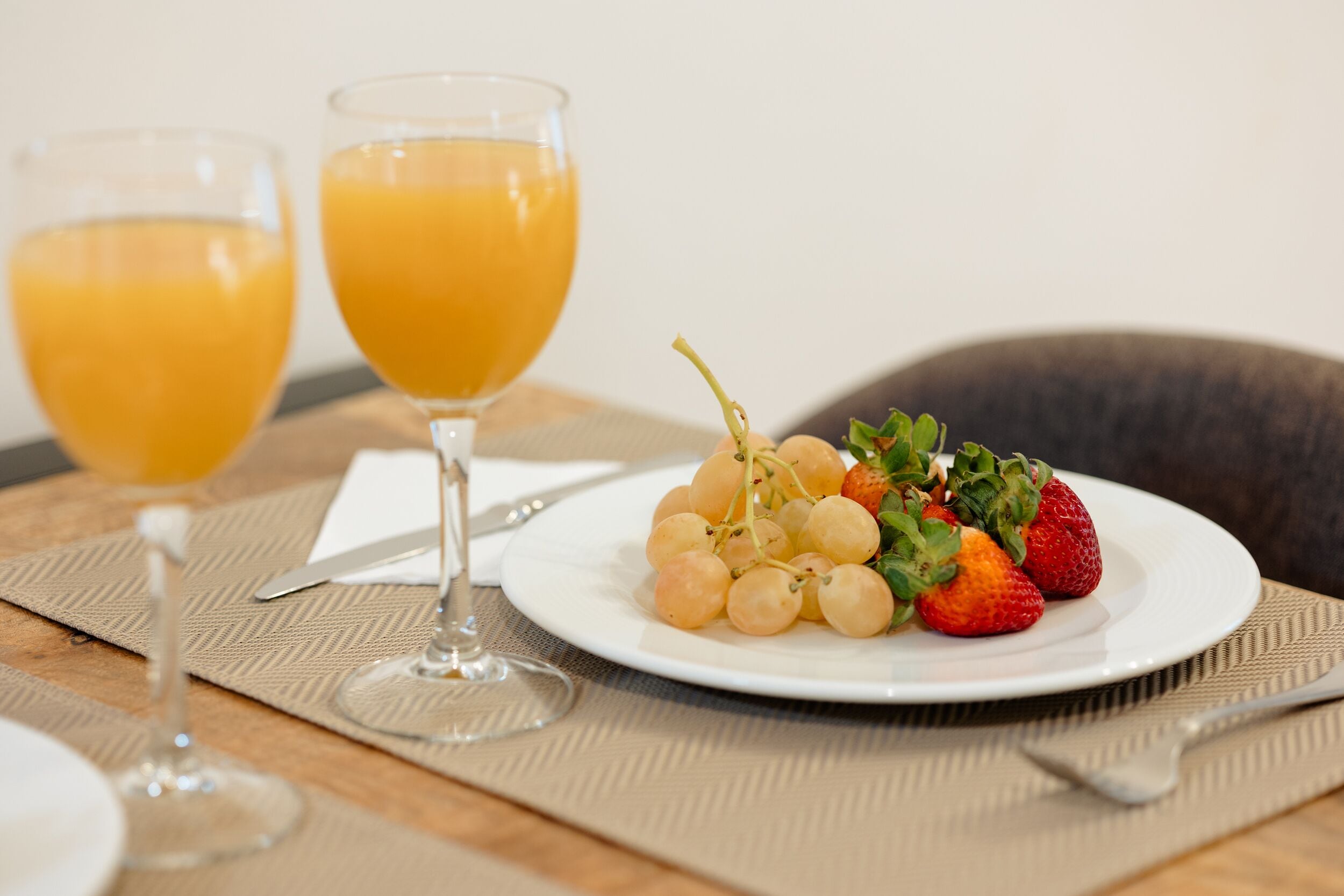 A breakfast setting with two glasses of orange juice, a plate of grapes and strawberries, and a fork and knife on a placemat.