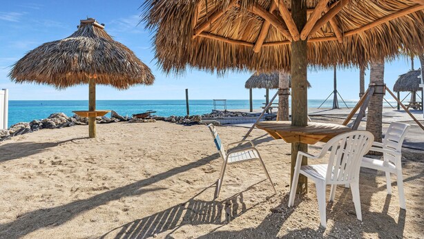 A beach with thatched umbrellas, white plastic chairs, and a wooden table. - 1 Island Hideaway in the Keys (Marathon)