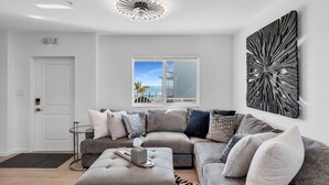 A modern living room with a grey sectional sofa, a black and white abstract wall art, a ceiling light fixture, and a window with a view of a building and palm trees. - 1 Island Hideaway in the Keys (Marathon)