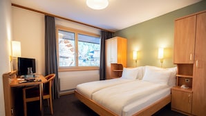 Een hotelkamer met een bed, bureau, stoel, kledingkast, bedlampjes en een raam met uitzicht op gebouwen.