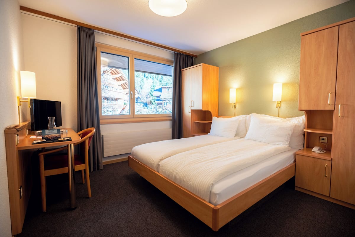 Habitación de hotel con cama, escritorio, silla, armario, lámparas de noche y ventana con vista a edificios.