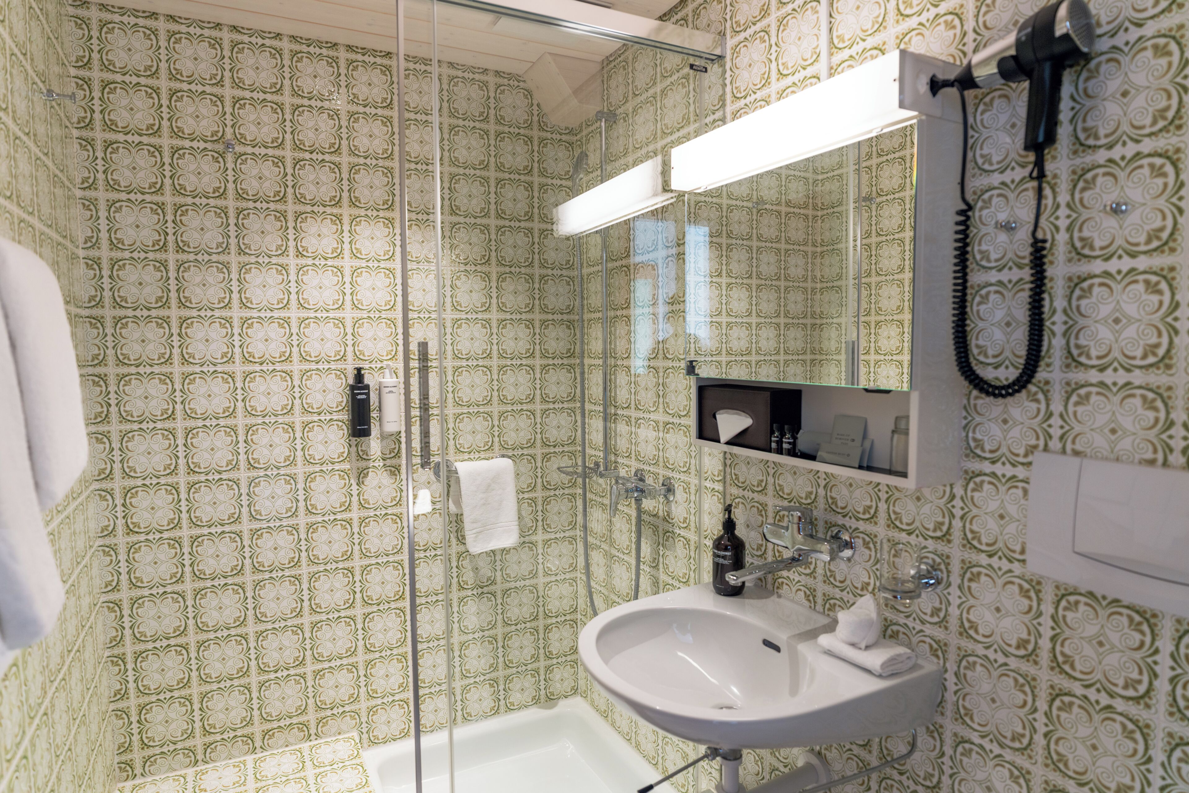 Un baño con papel tapiz estampado, cabina de ducha de vidrio, lavabo blanco y un espejo sobre el lavabo.