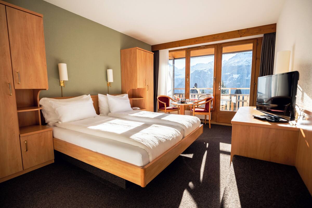 Habitación de hotel con una cama grande, muebles de madera, televisor y balcón con vistas a la montaña.