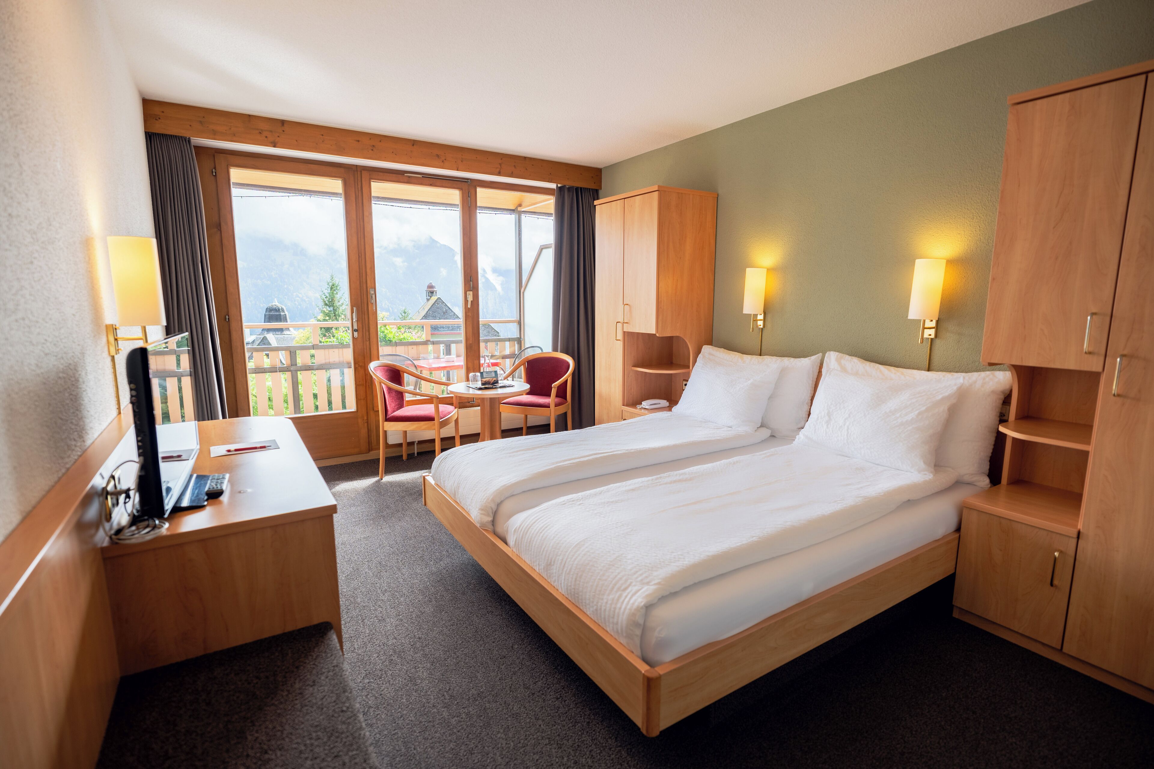 Une chambre d’hôtel avec un grand lit, une armoire en bois, un bureau avec un ordinateur et un balcon équipé de chaises et d’une table.