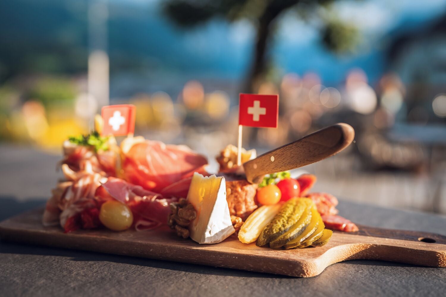 Tábua de madeira com diversos queijos, carnes e picles, acompanhada de uma faca e uma bandeira suíça.