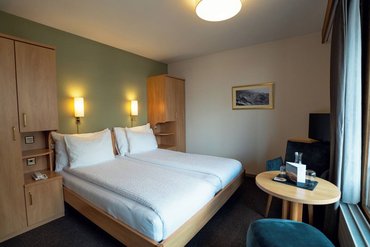 Habitación de hotel con cama, mesitas de noche, escritorio, silla, lámpara, un cuadro en la pared y una ventana con cortinas.
