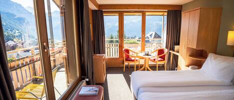 Hotelkamer met een groot bed, een bureau met stoel, een kleine tafel en een bankje. Er is een balkon met uitzicht op bergen en een stad.
