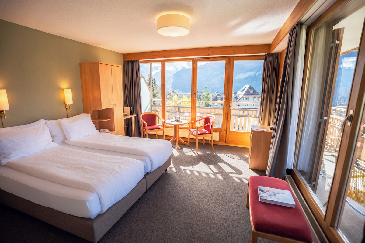 A hotel room with a large bed, a small table, two chairs, a bench, and a view of mountains through large windows.