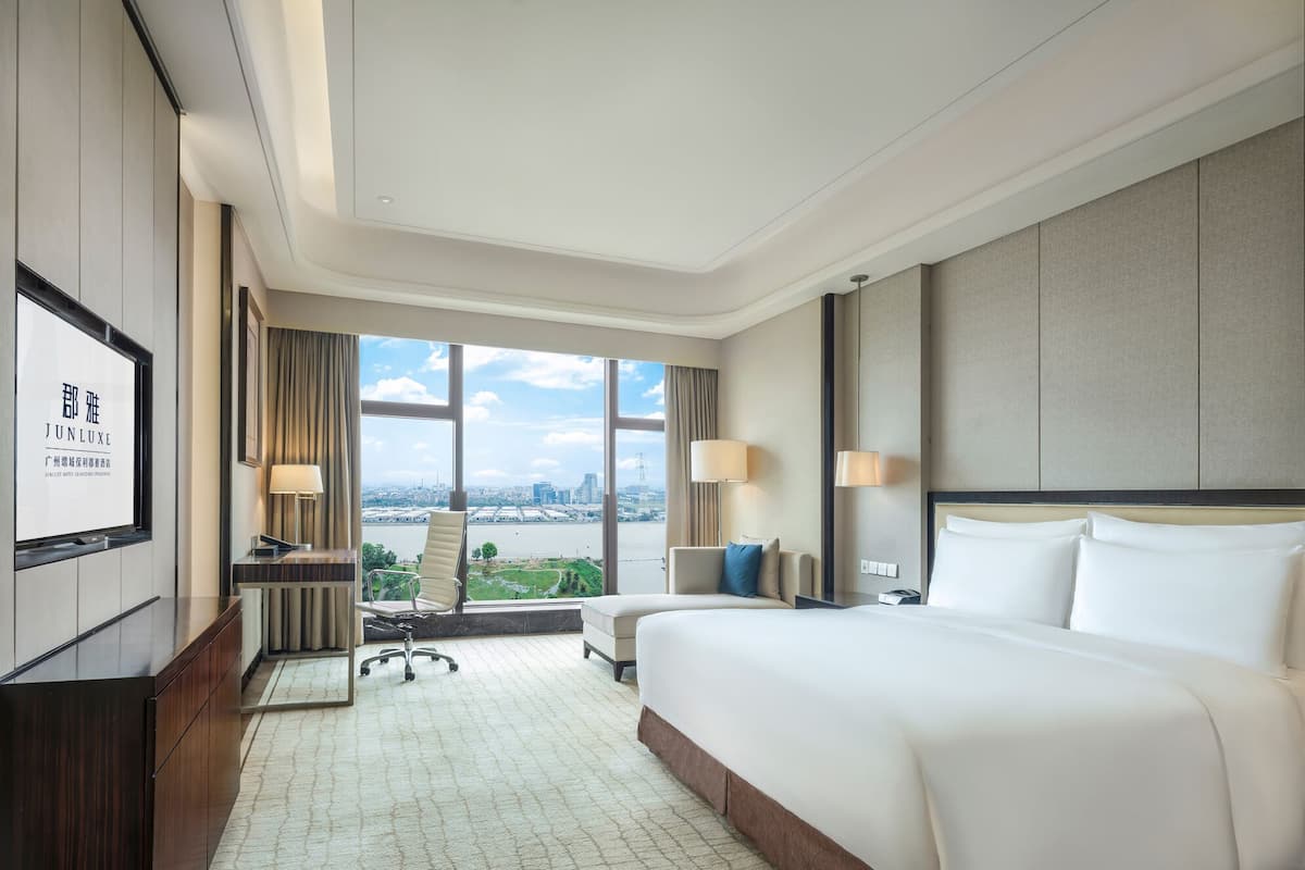A hotel room with a large bed, a desk, a chair, and a view of a cityscape through large windows.