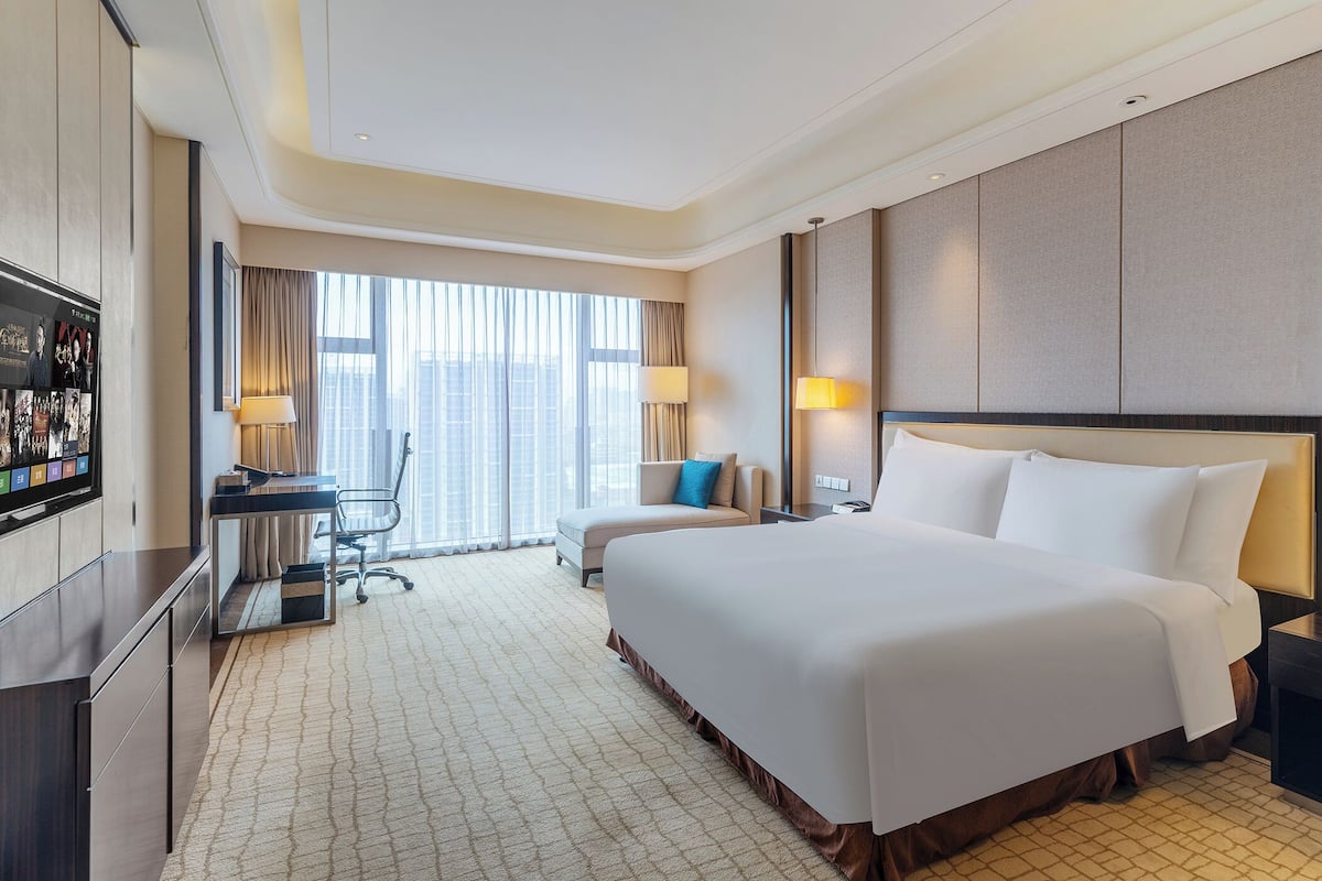 A hotel room with a large bed, a desk with a chair, a TV, and a view of the city through large windows.