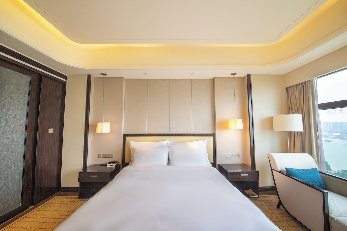 A hotel room with a large bed, two bedside tables, a chair, and a view of the city.