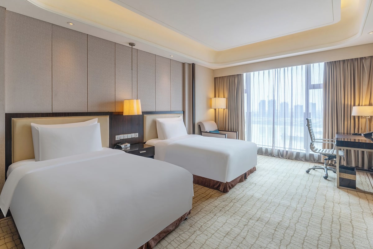 A hotel room with two beds, a desk, a chair, and a large window with curtains.