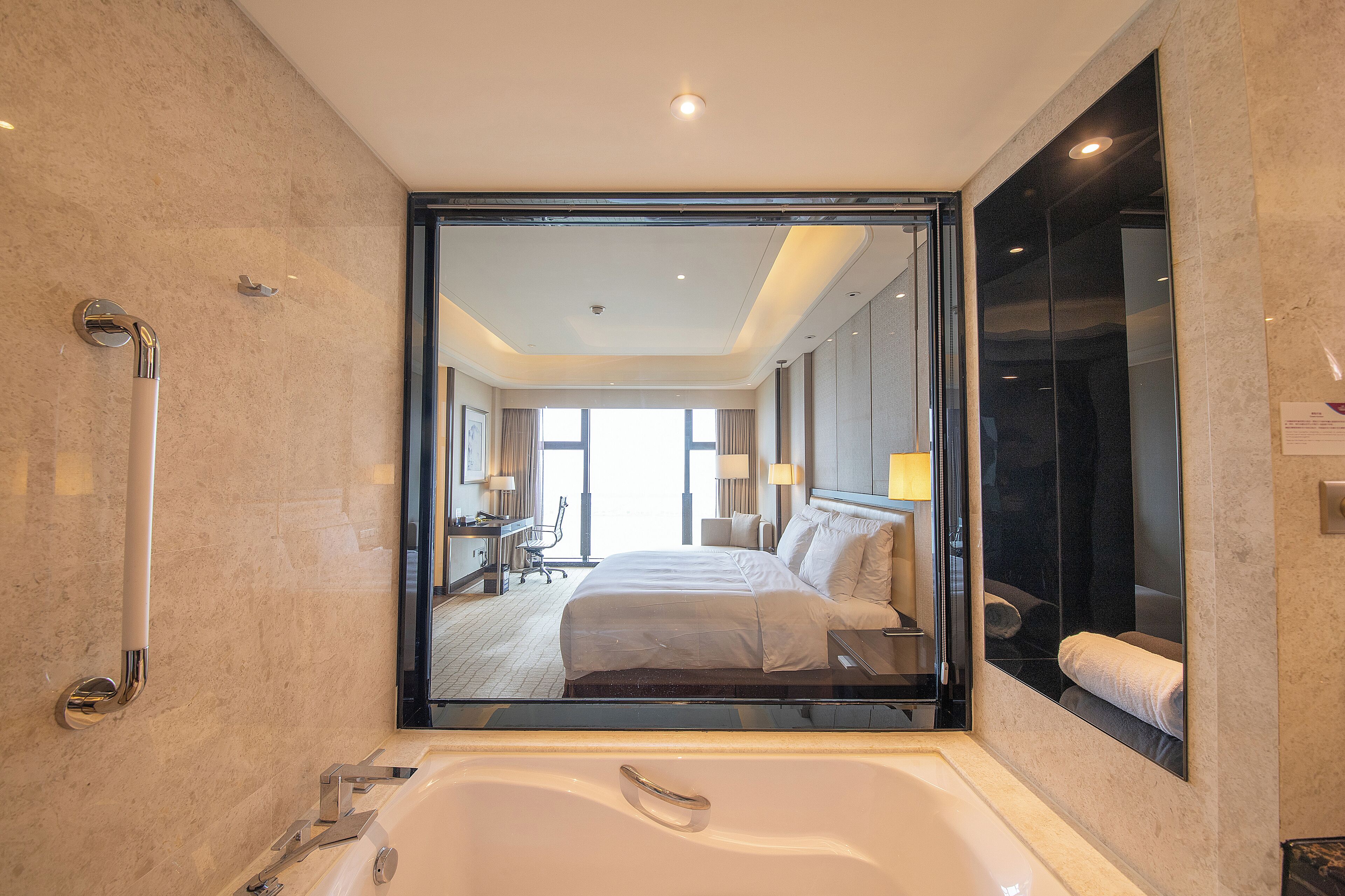 Un baño de hotel moderno con una bañera grande, una ducha y un espejo que refleja un dormitorio con cama y escritorio.