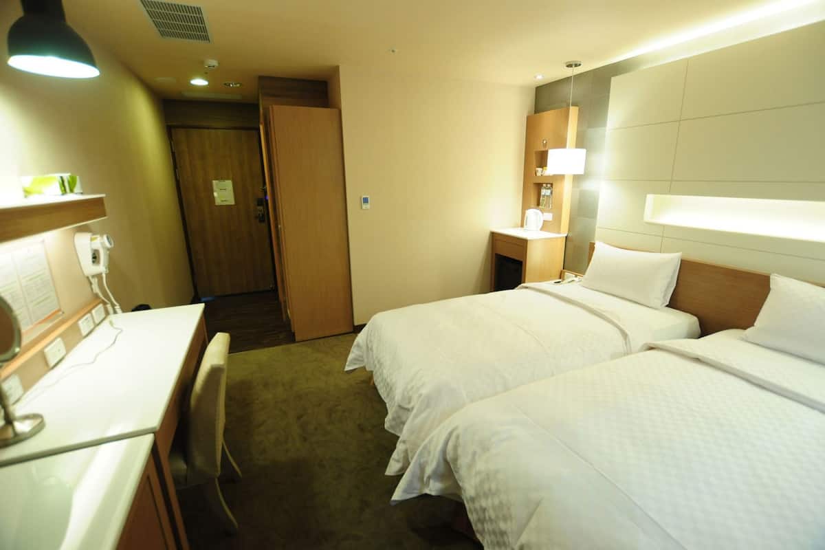 A hotel room with two beds, a desk, a chair, a nightstand, and a lamp.