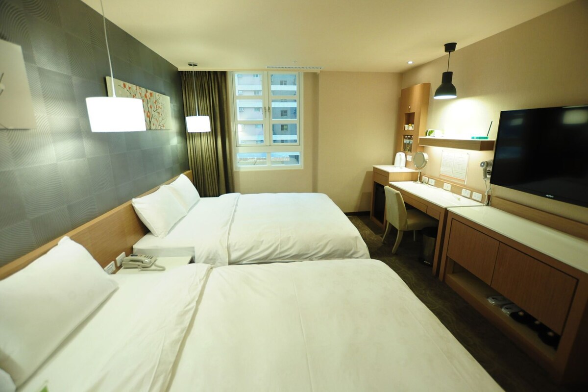 Habitación de hotel con dos camas, un escritorio con silla, televisor y ventana con cortinas.