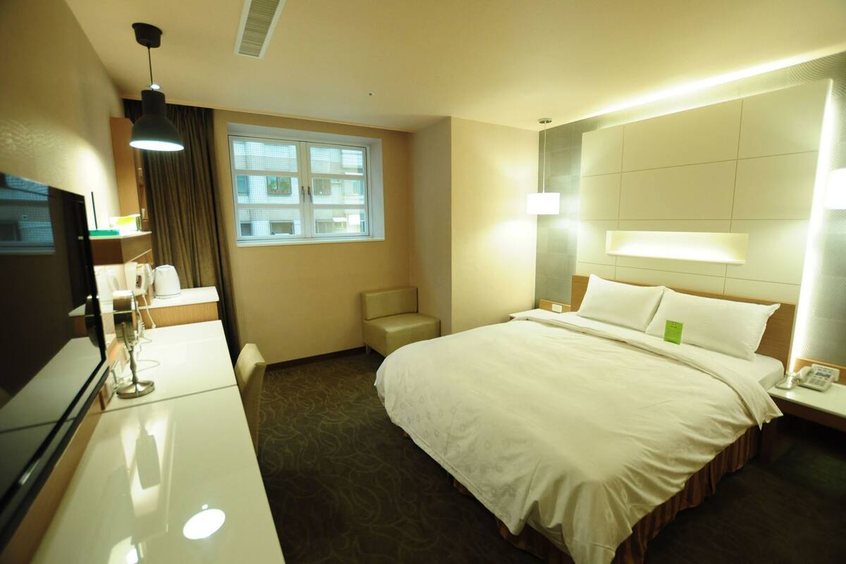 A hotel room with a bed, bedside table, lamp, TV, and a window.