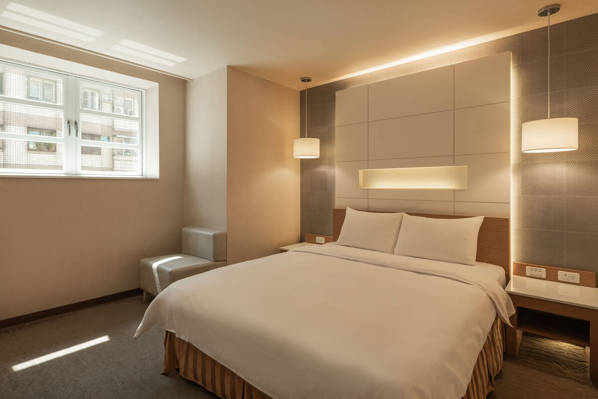 A hotel room with a large bed, bedside tables, a nightstand, a window with a view of buildings, and a wall-mounted light fixture.