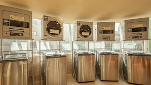 Una hilera de lavadoras de acero inoxidable con paneles de control, ubicadas en una habitación bien iluminada con ventanales grandes.