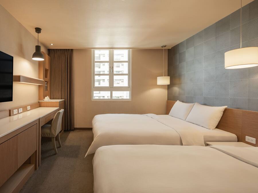 Quarto de hotel com duas camas, uma escrivaninha, uma cadeira, televisão e janela com vista para edifícios.