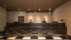 Un área de recepción con mostrador de mármol, dos recepcionistas e iluminación colgante.