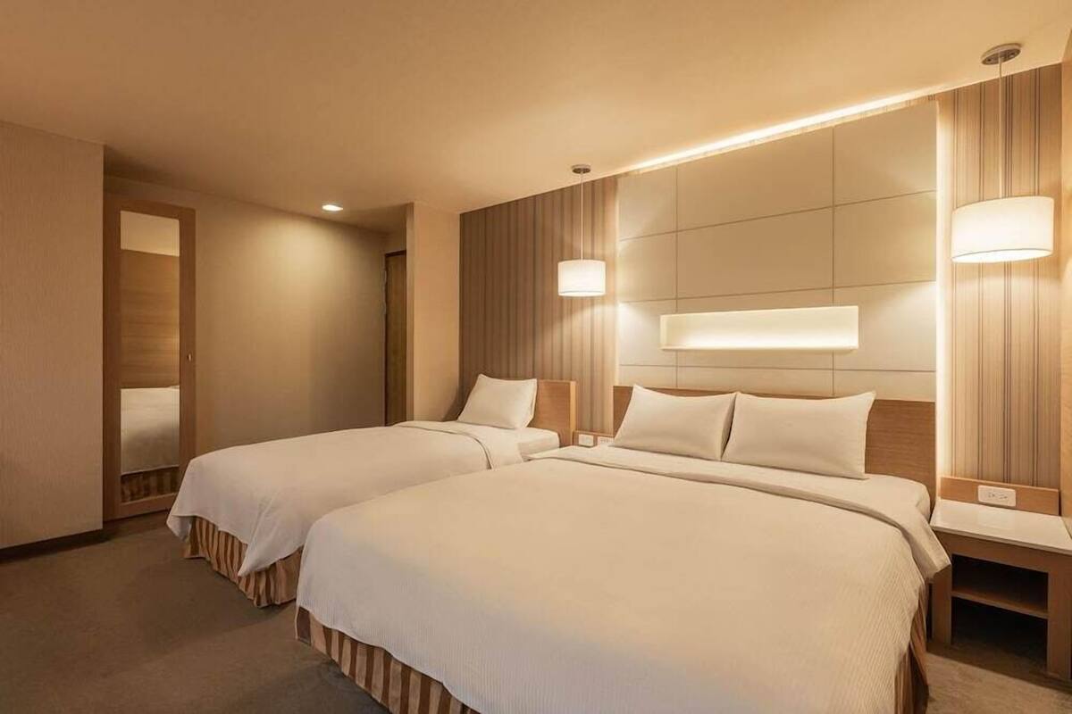 A hotel room with two beds, a wooden headboard, bedside lamps, and a carpeted floor.