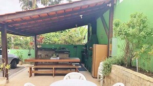 Un patio al aire libre con una mesa de madera y sillas de plástico blanco, una pared verde y una zona cubierta con una parrilla.