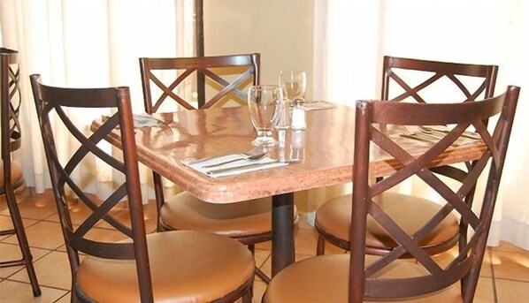 Un comedor con una mesa de vidrio, cuatro sillas de madera con asientos acolchados, y cubiertos y vasos sobre la mesa.