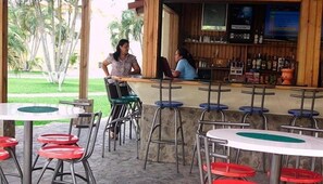 An outdoor cafe with tables and chairs, a bar area with bottles, and a view of greenery.