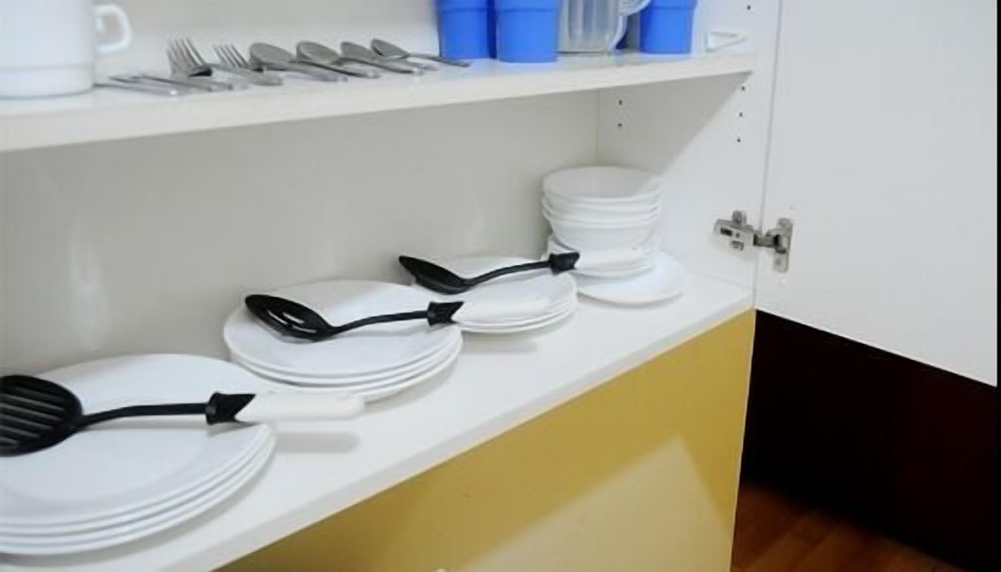 Una cocina con gabinetes blancos y amarillos, una encimera blanca y diversos utensilios y platos de cocina ordenados en las estanterías.