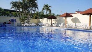 Une piscine entourée de chaises longues et de parasols, bordée de palmiers et d’un bâtiment blanc.
