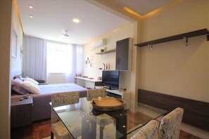 A modern hotel room with a bed, a TV, a dining area with a glass table, and a sofa.