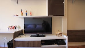 A modern living room with a flat-screen TV on a wooden entertainment unit, a white shelf with figurines, and a dark brown accent wall. - Assis 801 (Rio de Janeiro)